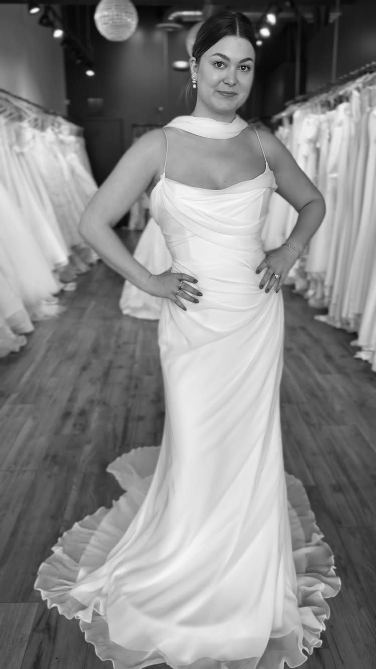 A woman posing in a white sleeveless wedding gown with a draped neckline and flowing train.