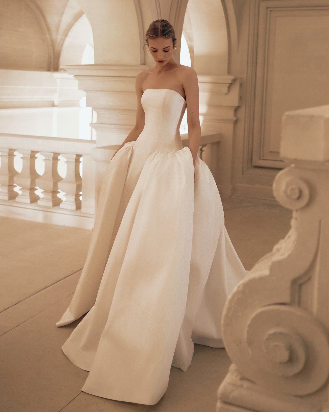 Woman wearing a strapless white Mikado wedding gown with a voluminous skirt in a classical stone building.