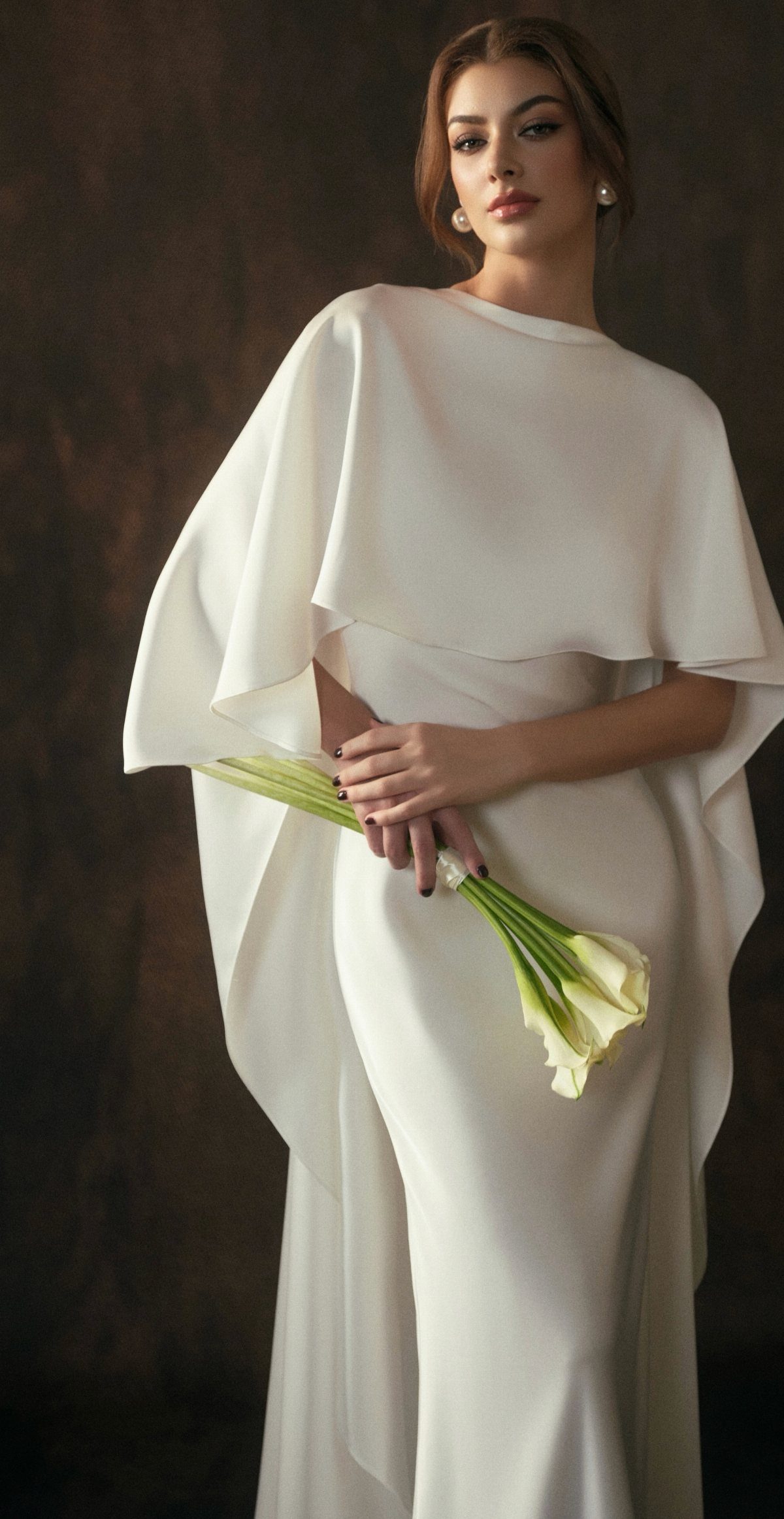 A woman in a white minimalist wedding gown holding white calla lilies against a dark background.