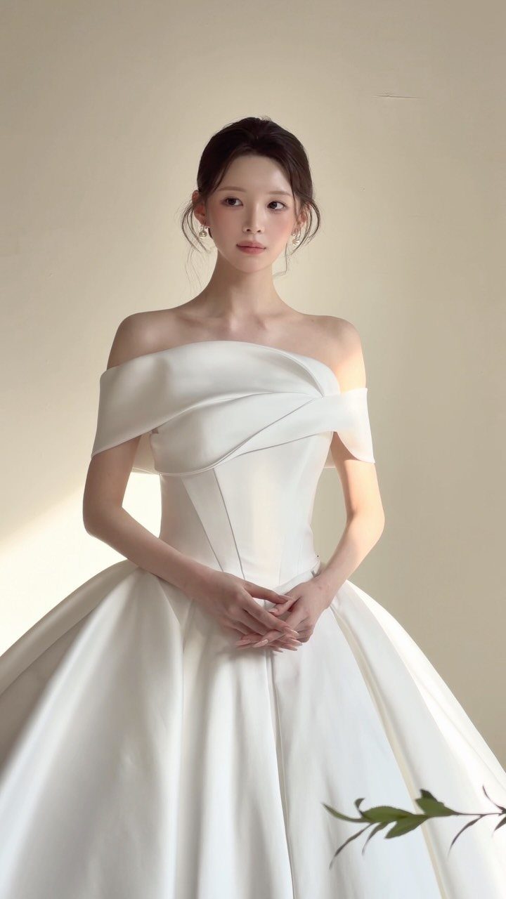 A woman wearing a minimalist off-the-shoulder white satin wedding gown against a neutral background.