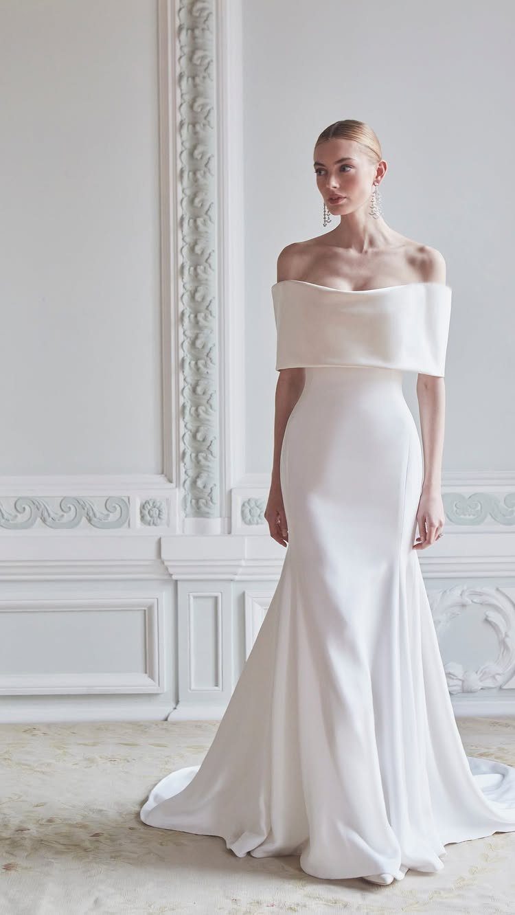 Woman wearing a white off-the-shoulder minimalist wedding gown standing in a white room.