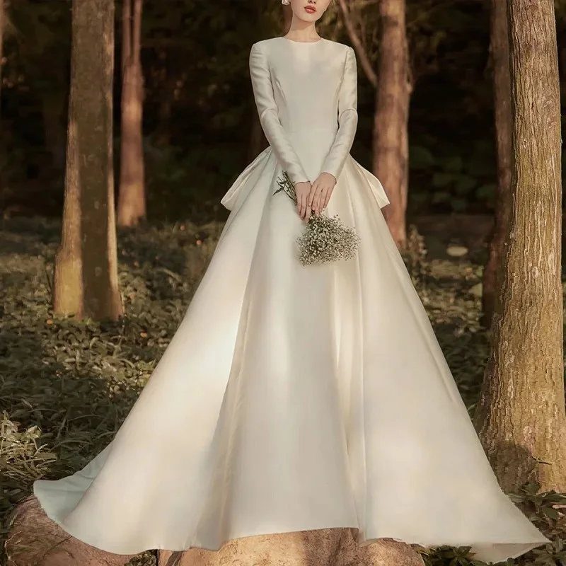 Woman in a long-sleeved white silk wedding gown holding flowers in a forest.