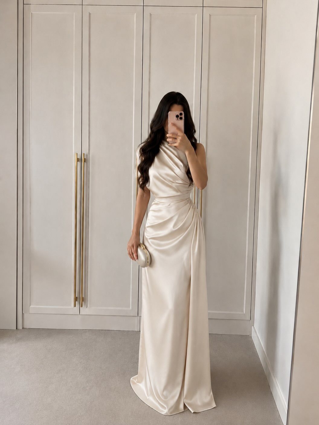 Woman in a draped cream silk satin one-shoulder evening dress holding a clutch.