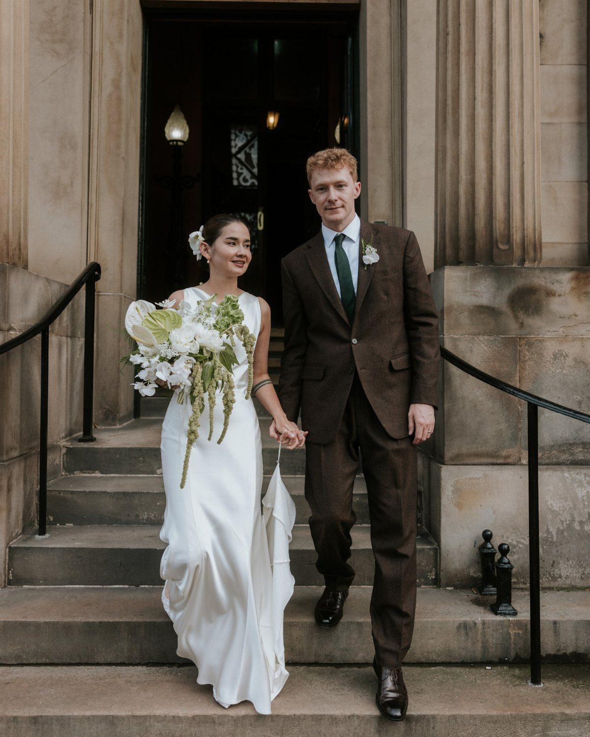 A bride in a white satin gown and a groom in a brown suit walk down stone steps.