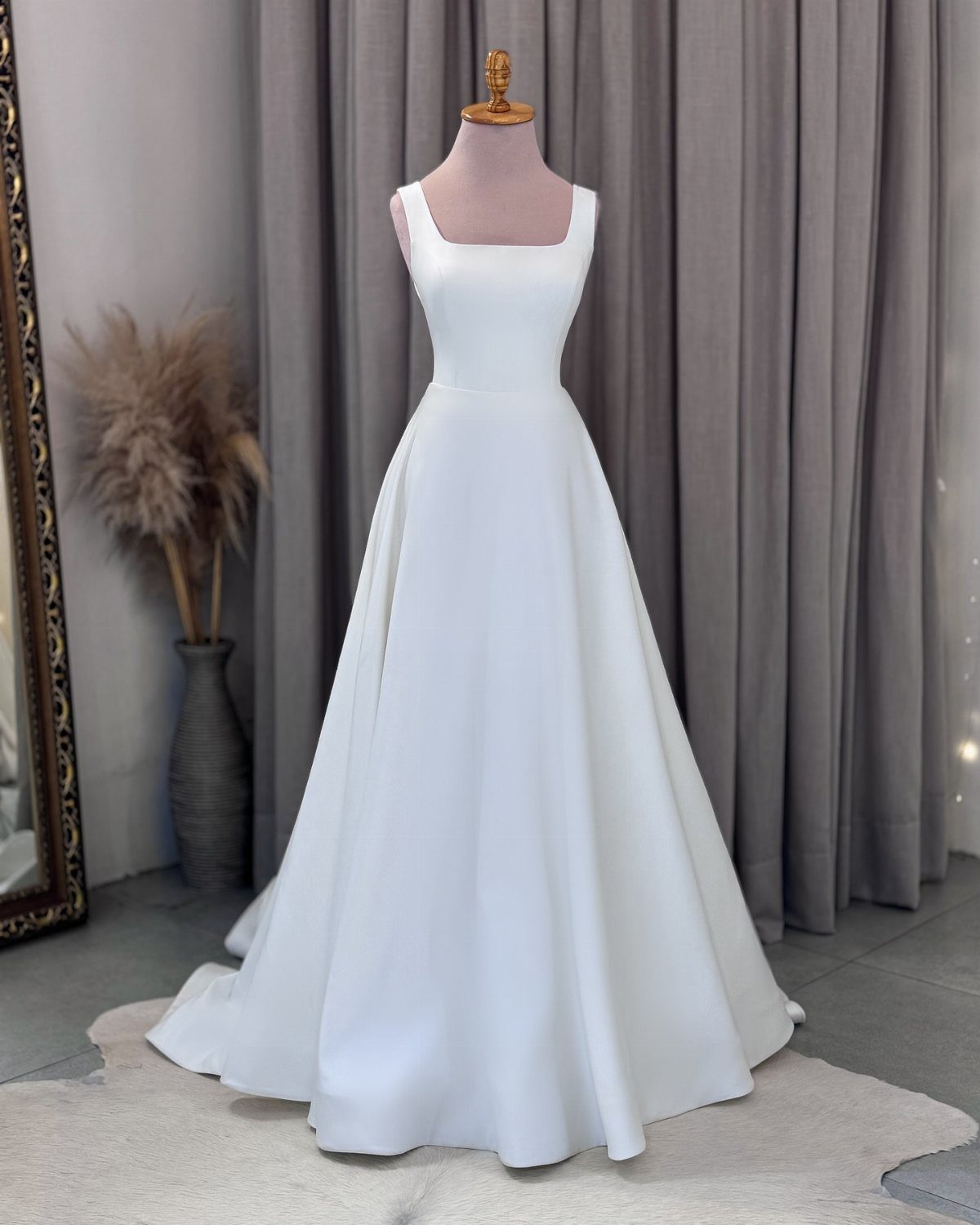 A white minimalist wedding gown with a square neckline displayed on a mannequin against grey curtains.
