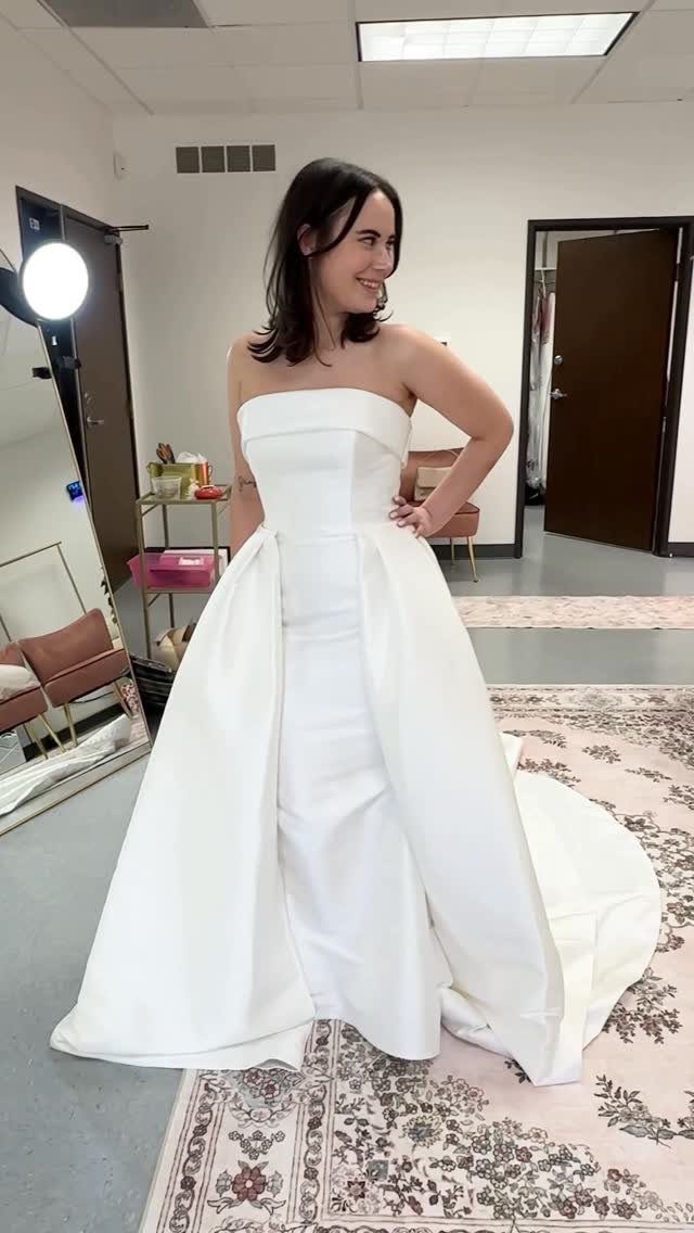 A smiling woman wearing a strapless white minimalist wedding gown with a voluminous overskirt.