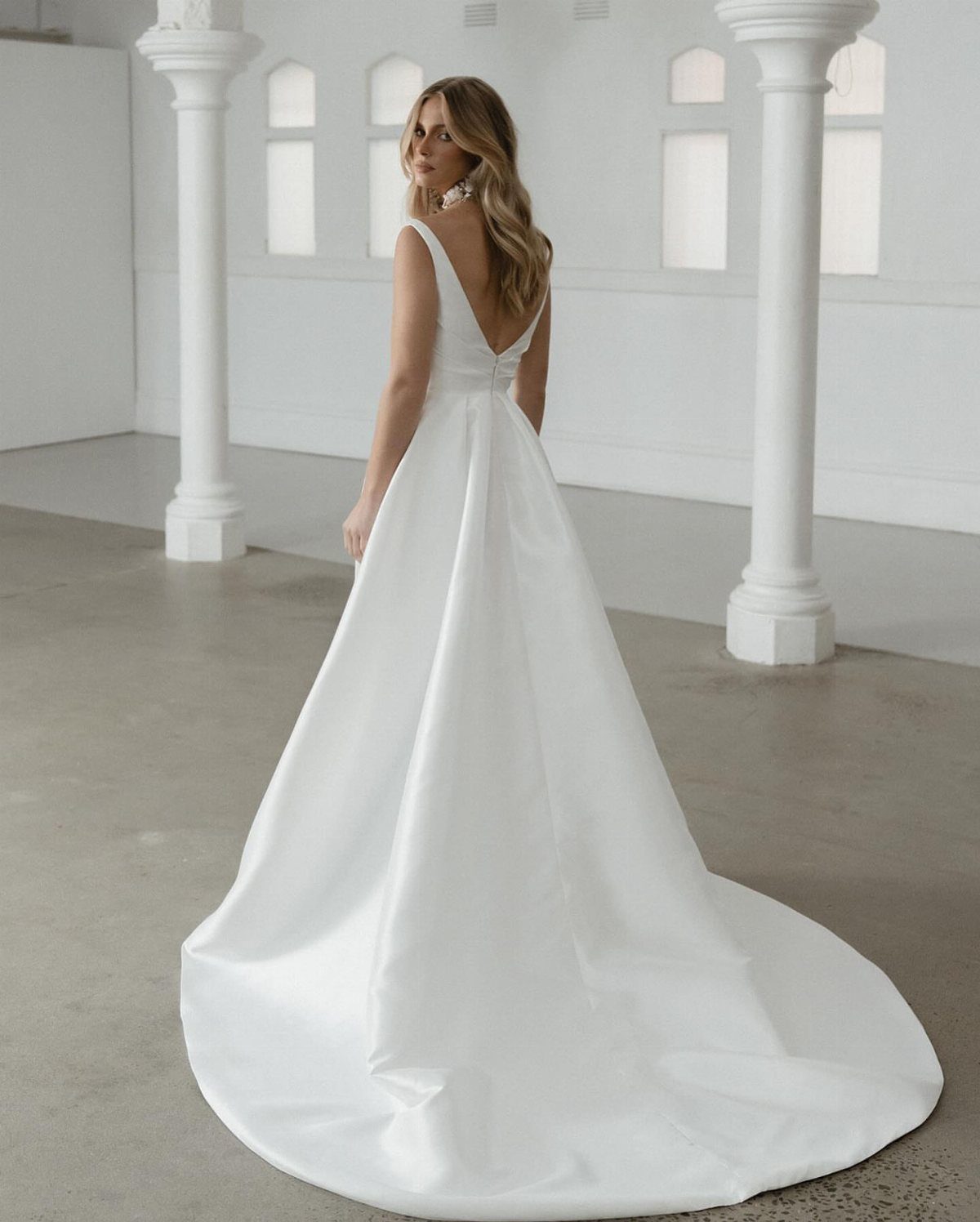 Woman in a white satin wedding gown with a long train standing in a white room.