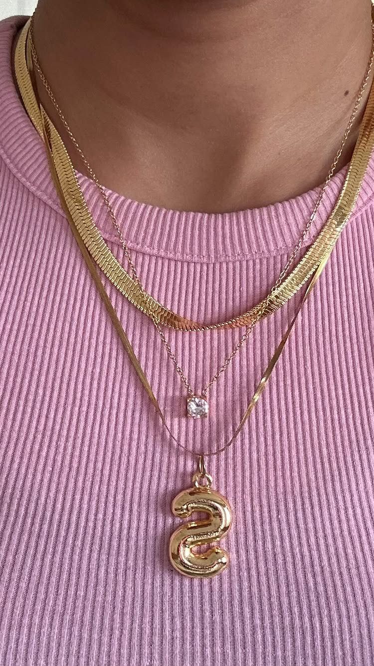 Three layered gold necklaces with different pendants worn over a pink ribbed shirt.