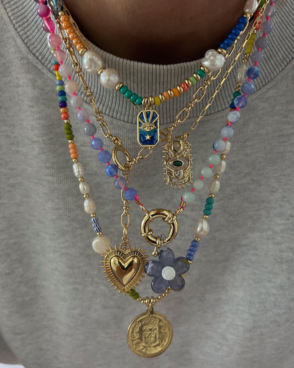 Layered necklaces with colorful beads and various gold charms like a heart and flower on a person.
