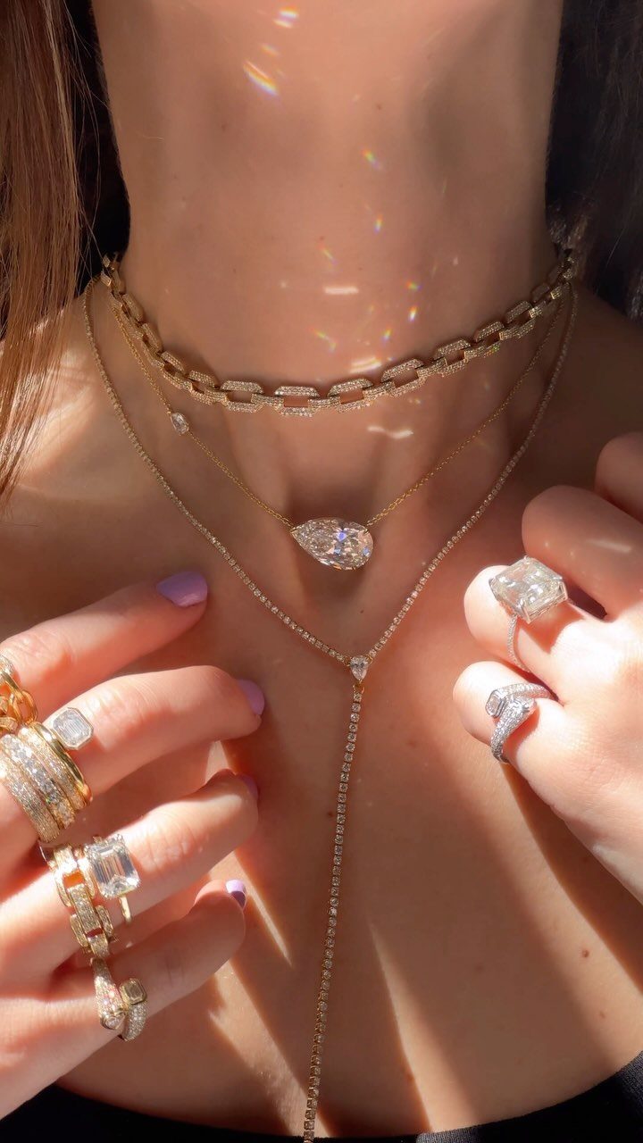 A person wearing layered gold necklaces with a large pear-shaped diamond pendant and multiple rings.