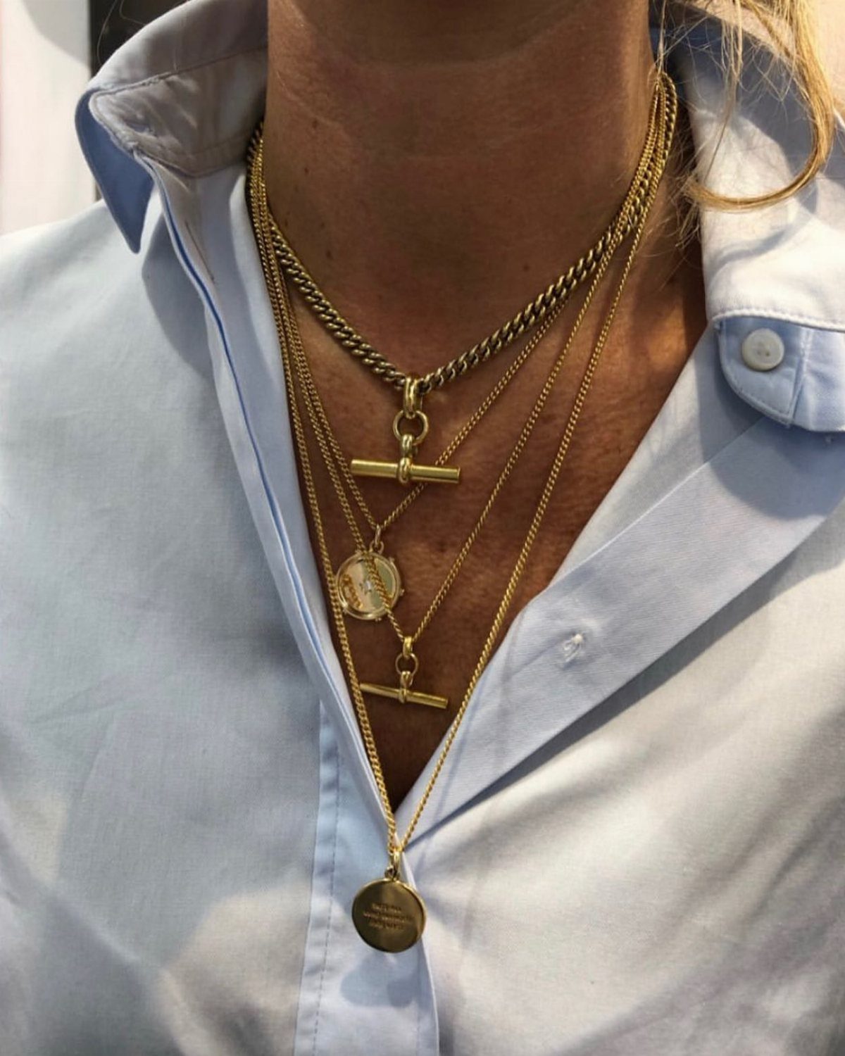 Multiple layered gold necklaces of varying thicknesses and textures worn over a light blue shirt.