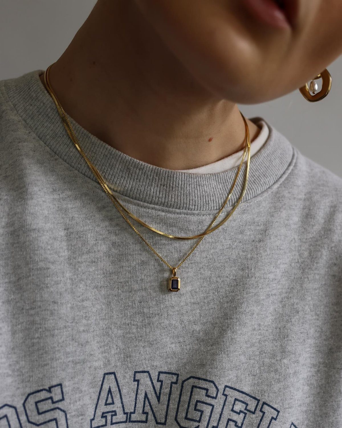 Two layered gold necklaces with a small pendant worn over a grey sweatshirt.