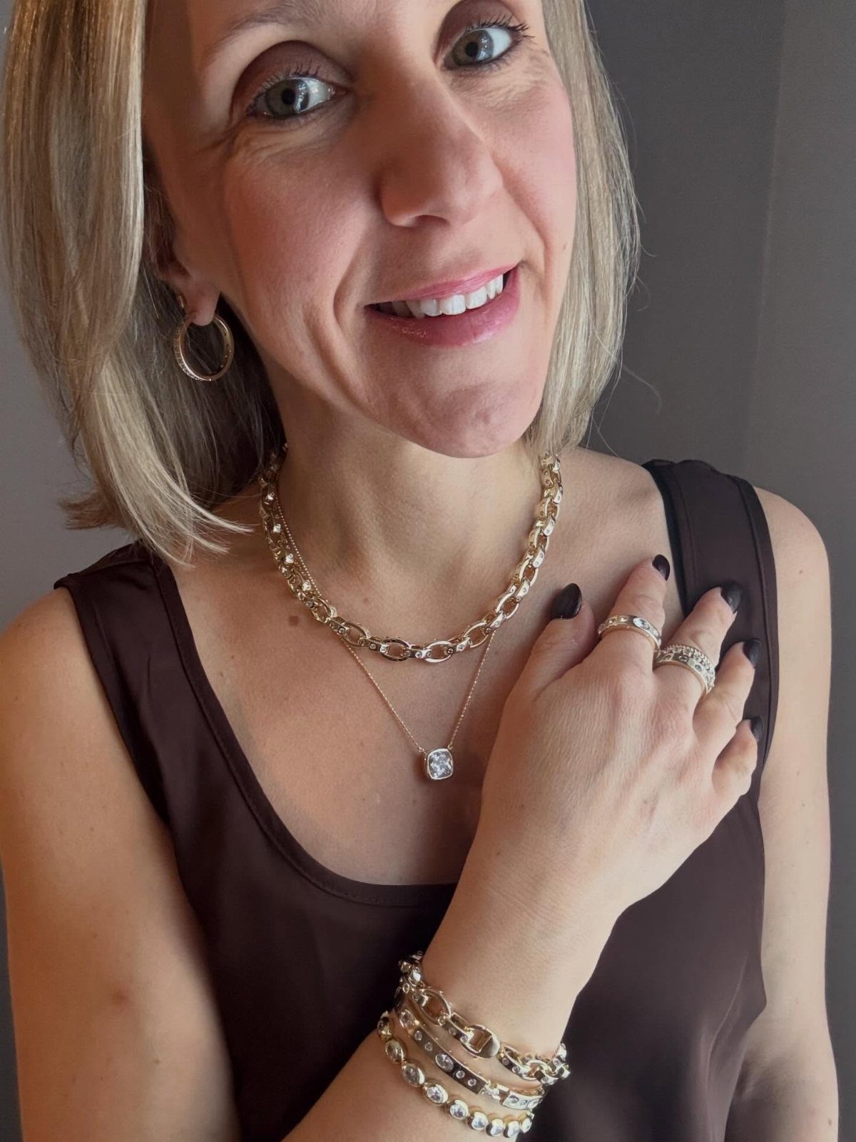 A smiling woman wearing layered gold necklaces and chunky gold bracelets against a plain background.