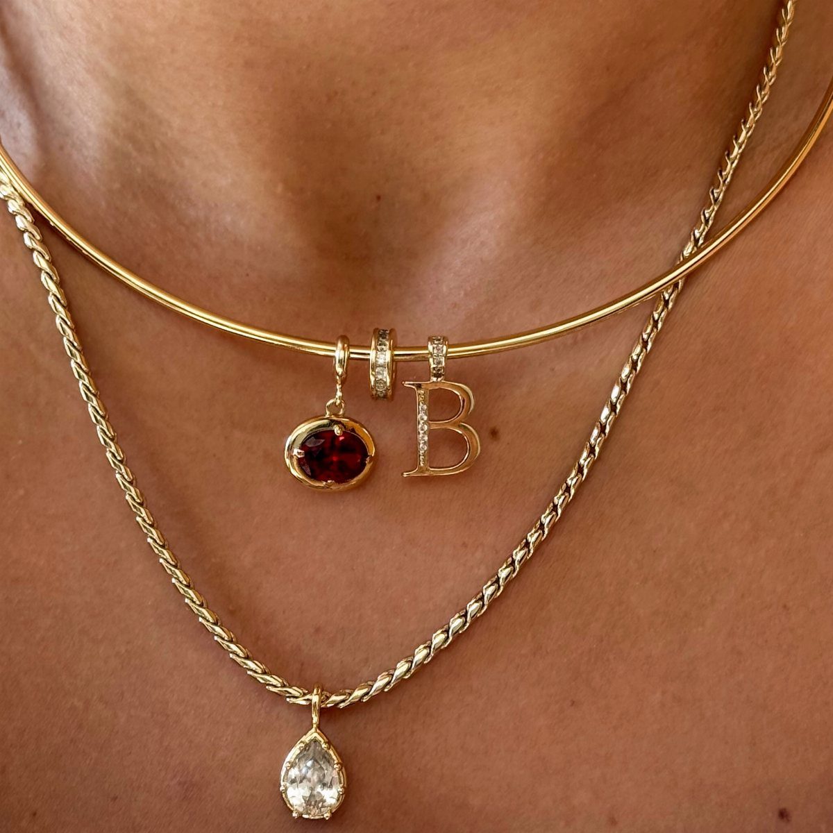 Three layered gold necklaces with a red gemstone, a letter B charm, and a pear-shaped pendant.