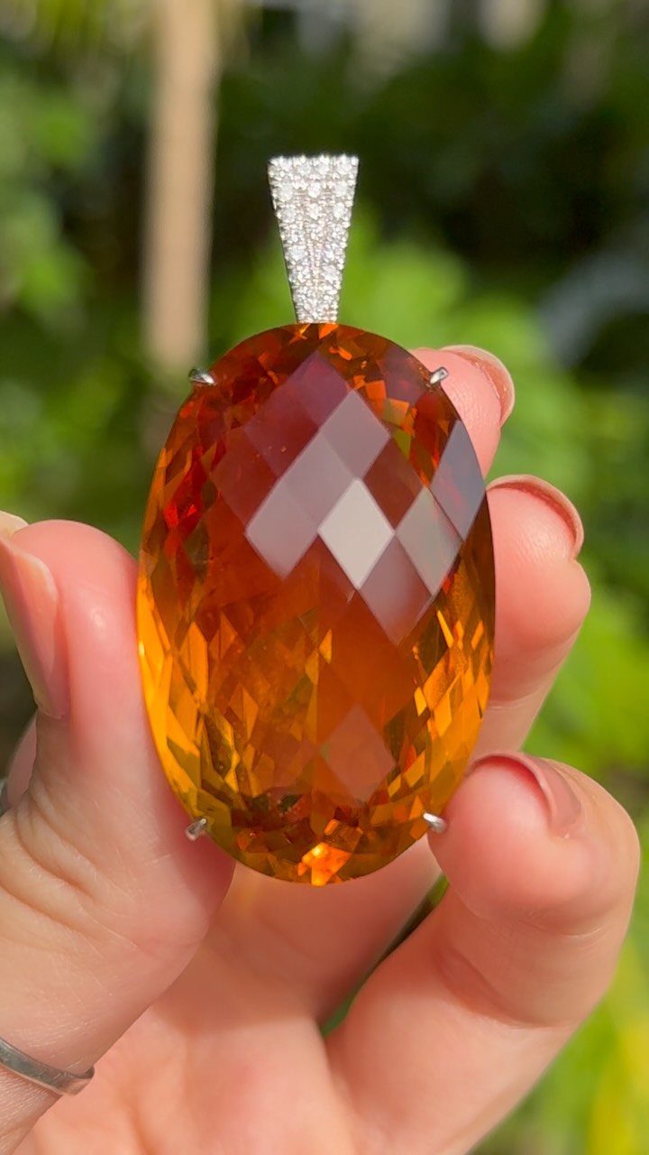 Precious Stones Jewelry: 11 Ideas From Emerald to Gold Bangles 3 A hand holding a large, faceted, oval orange citrine gemstone pendant against a blurred green background.