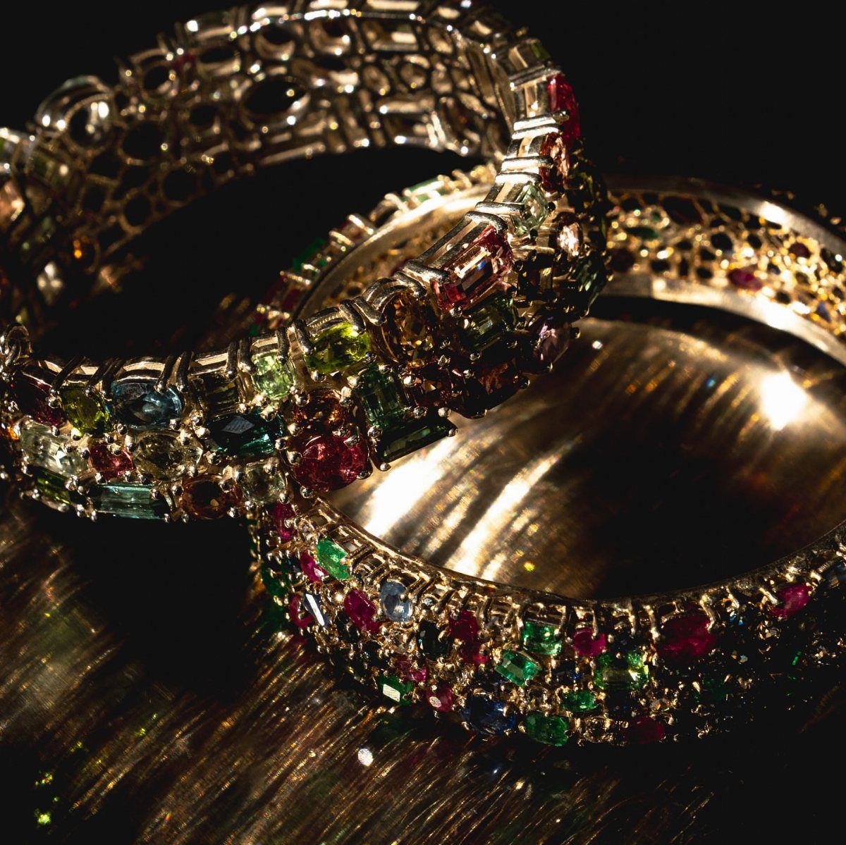 Precious Stones Jewelry: 11 Ideas From Emerald to Gold Bangles 11 Three gold bangles encrusted with colorful precious stones resting on a dark reflective surface.