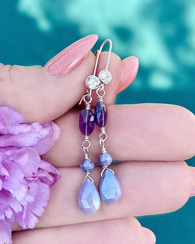 Precious Stones Jewelry: 11 Ideas From Emerald to Gold Bangles 9 A hand holding silver earrings with purple and blue faceted gemstones against a turquoise background.