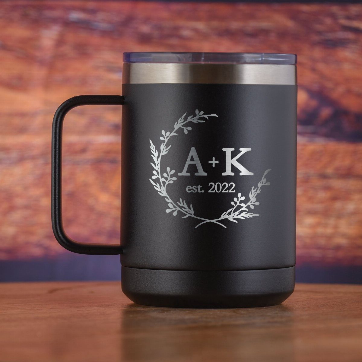 Rehearsal Dinner Gift Ideas: 9 Ways to Thank Your People 1 A matte black engraved travel mug with a floral wreath and initials on a wooden table.