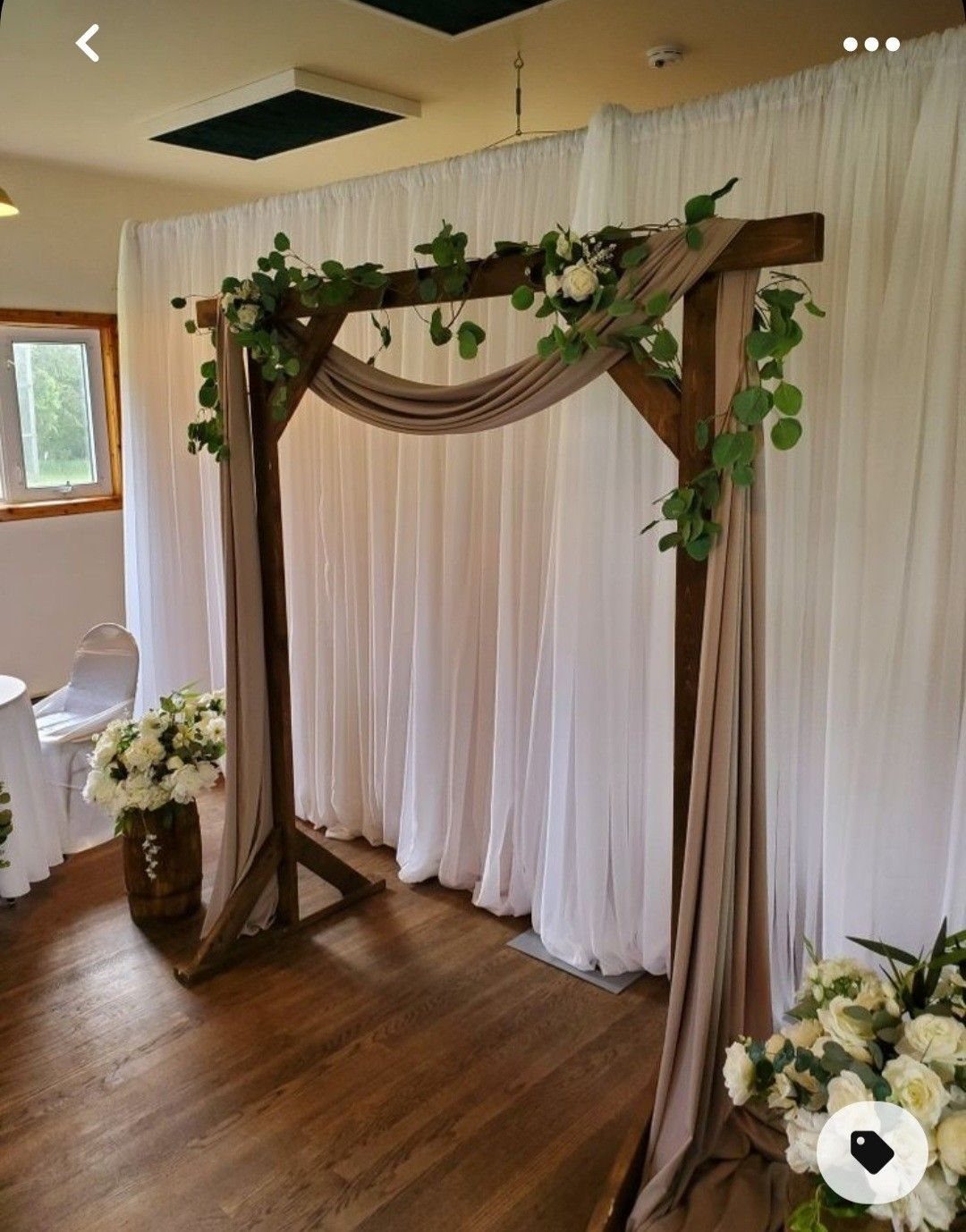 Wooden wedding arch with tan drapes and green floral vines against white curtains.