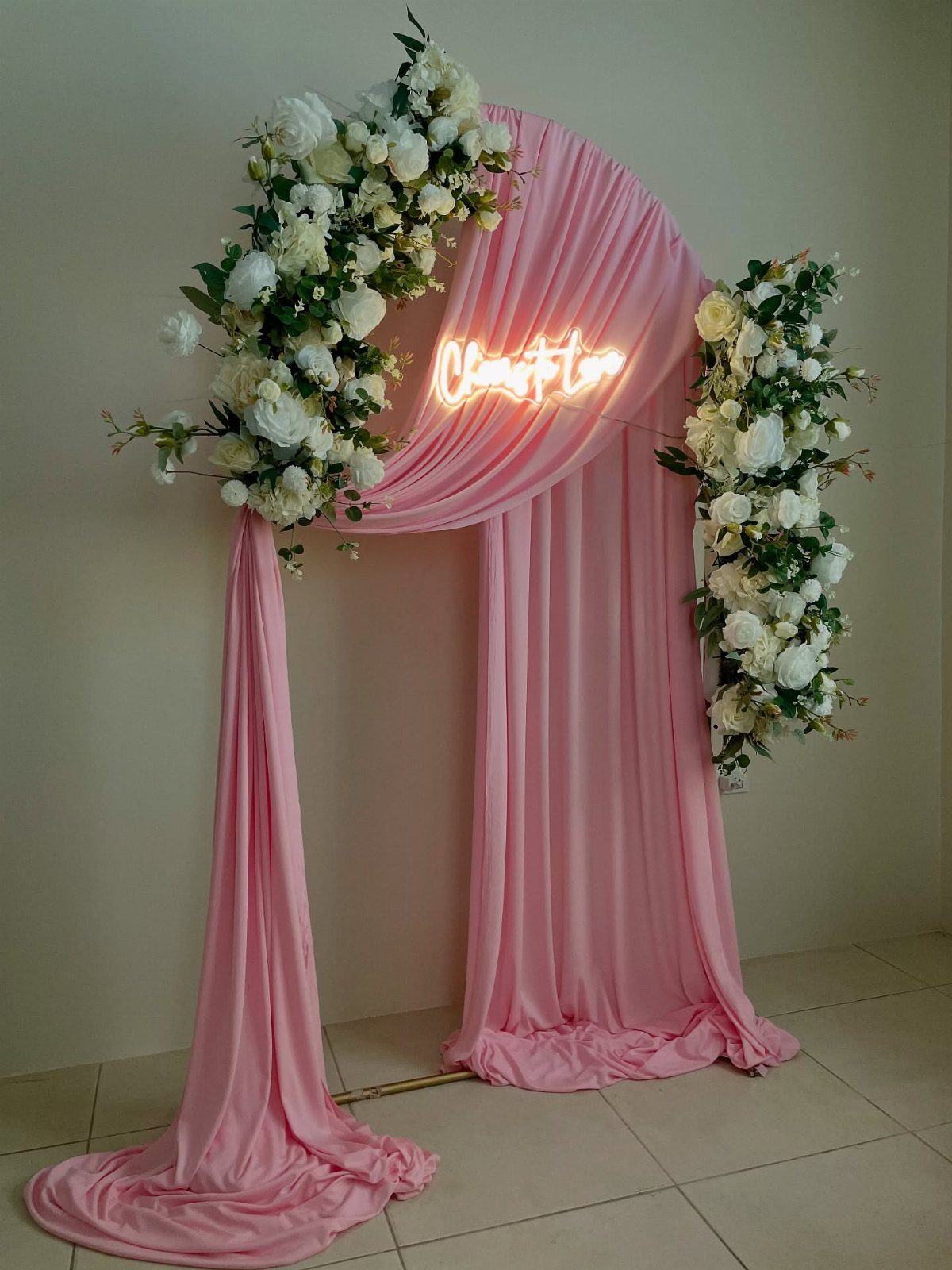 Pink draped wedding arch decorated with white roses and greenery featuring a neon sign.