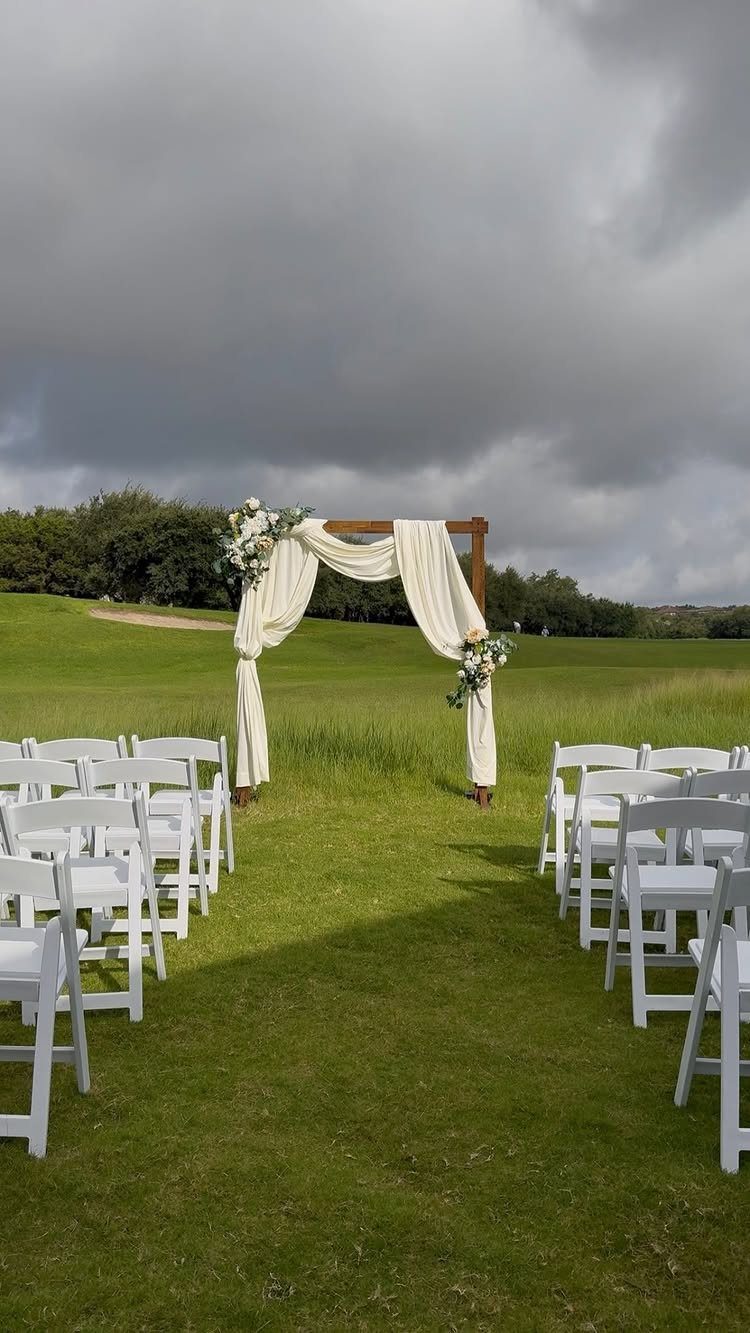A wooden wedding arch with white fabric drapes and flowers on a green lawn.