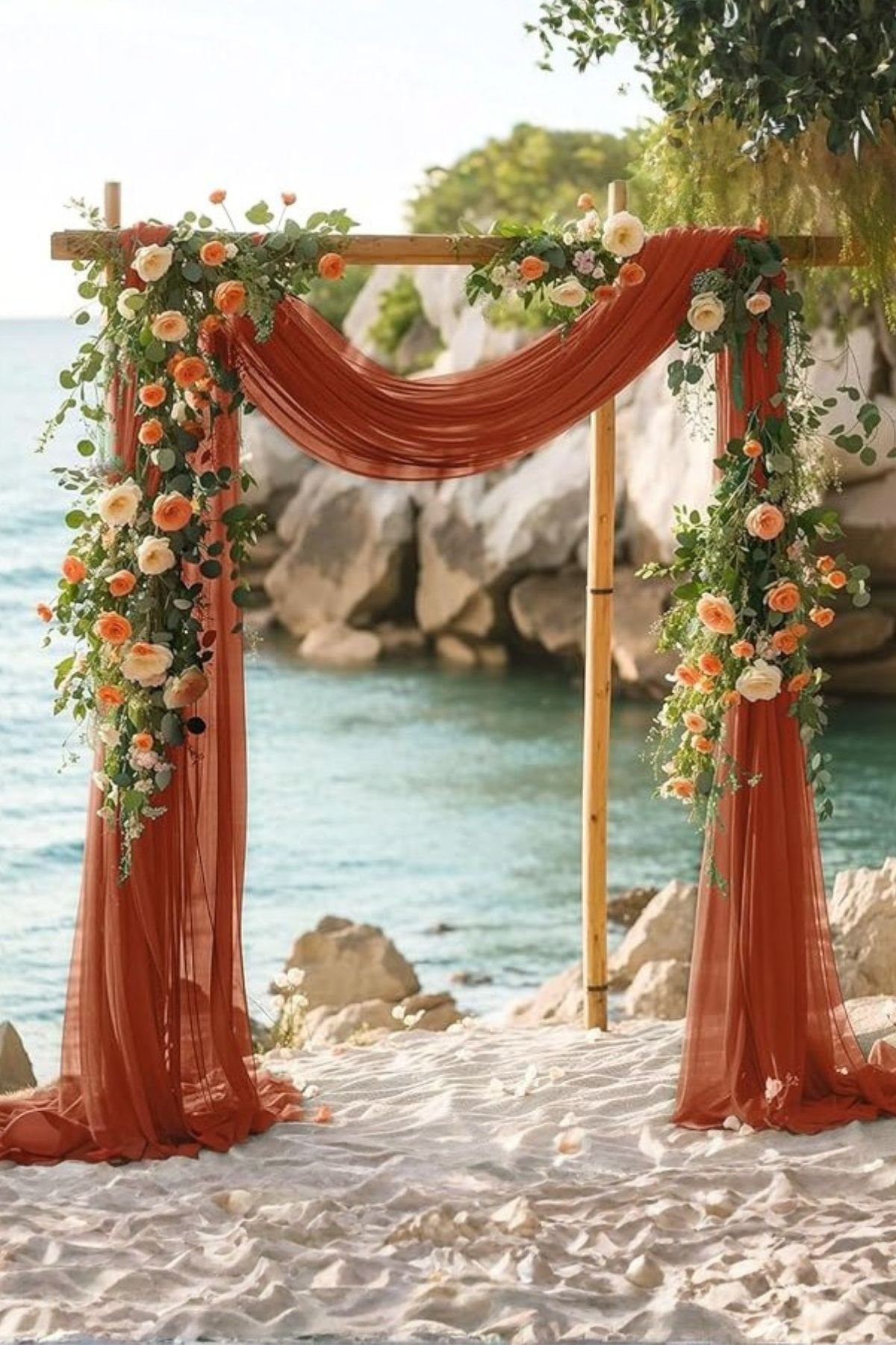 A wooden wedding arch with coral drapes and orange flowers on a sandy beach.