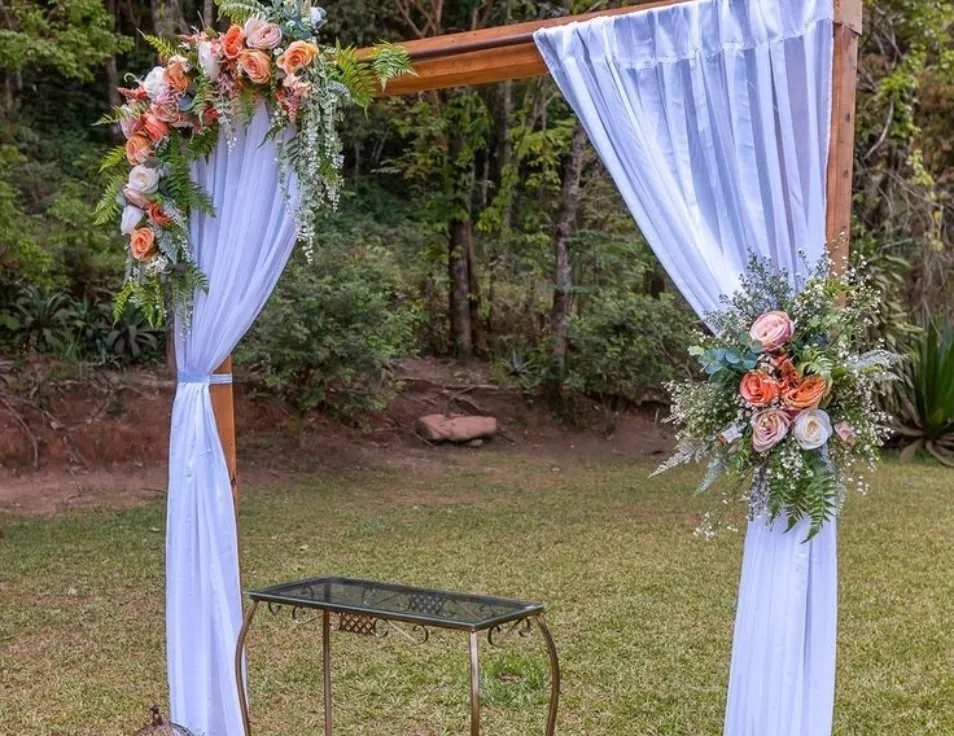 Wooden wedding arch with white drapes and peach floral arrangements on a green lawn.