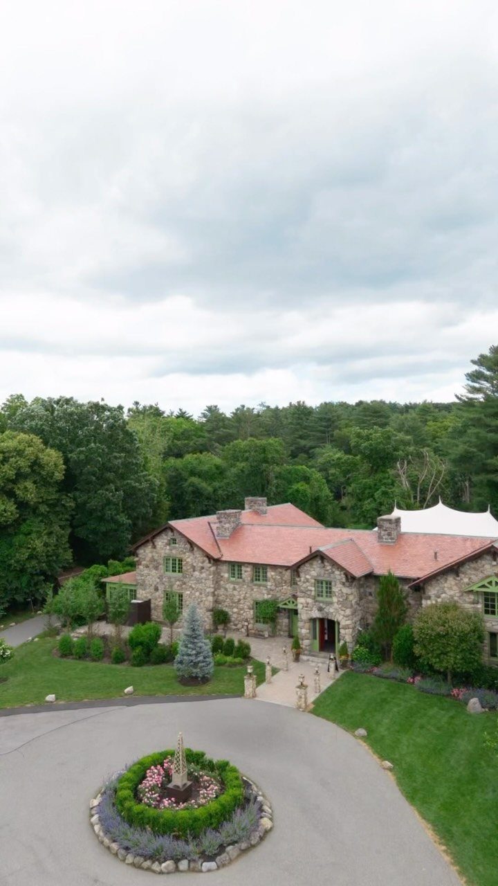 A large stone manor with a red roof surrounded by dense green forest and manicured lawns.