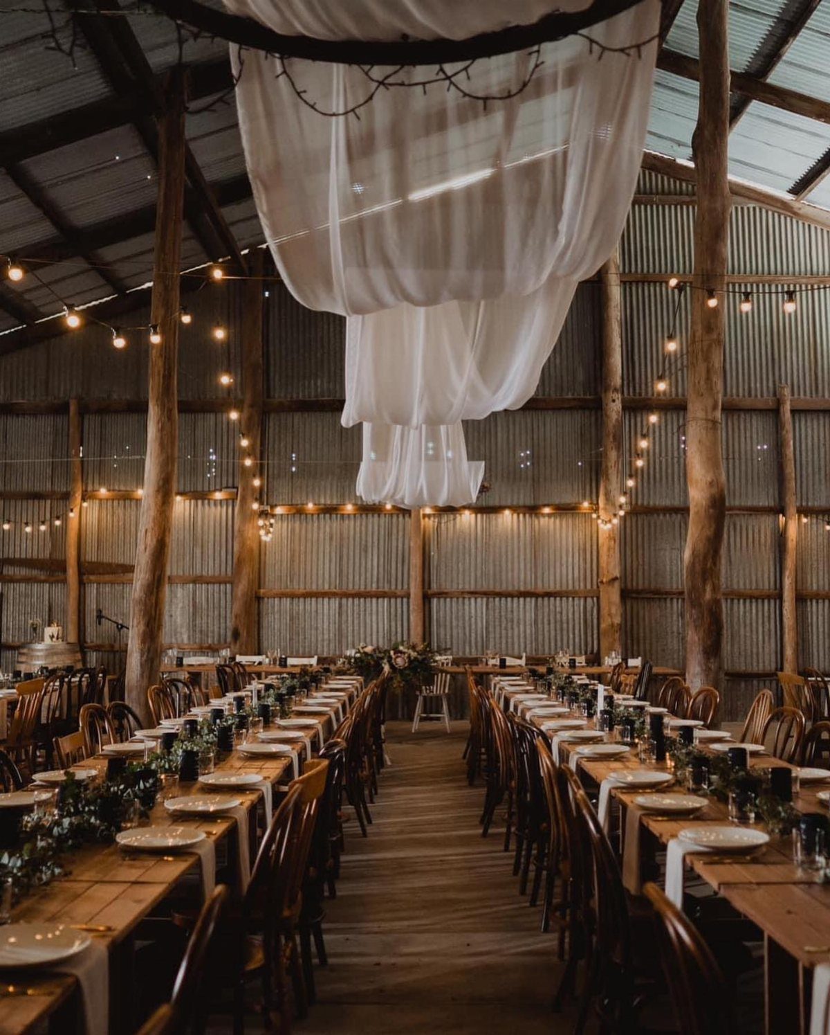 Long wooden dining tables set for a wedding inside a rustic corrugated metal barn.