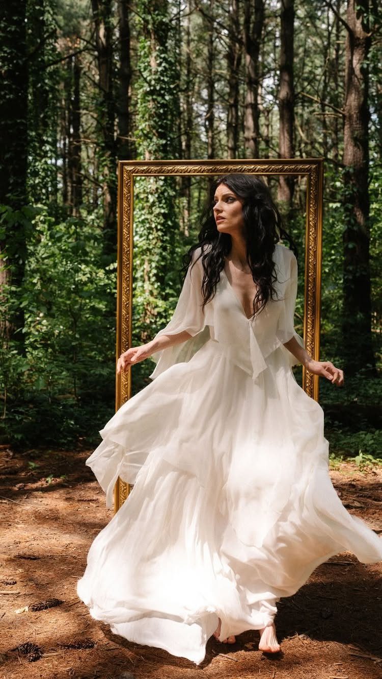 A woman in a flowing white chiffon wedding dress posing in a forest with a gold frame.