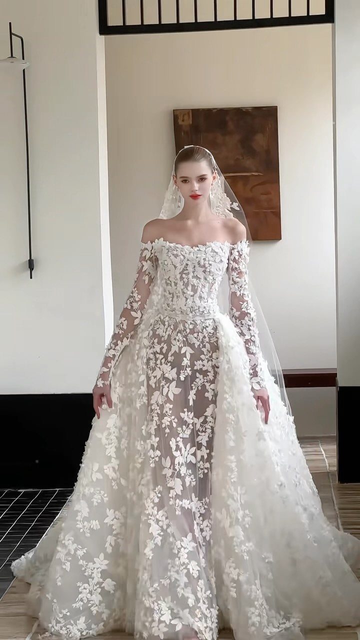 Woman wearing an off-the-shoulder white wedding gown with floral lace and a long veil.