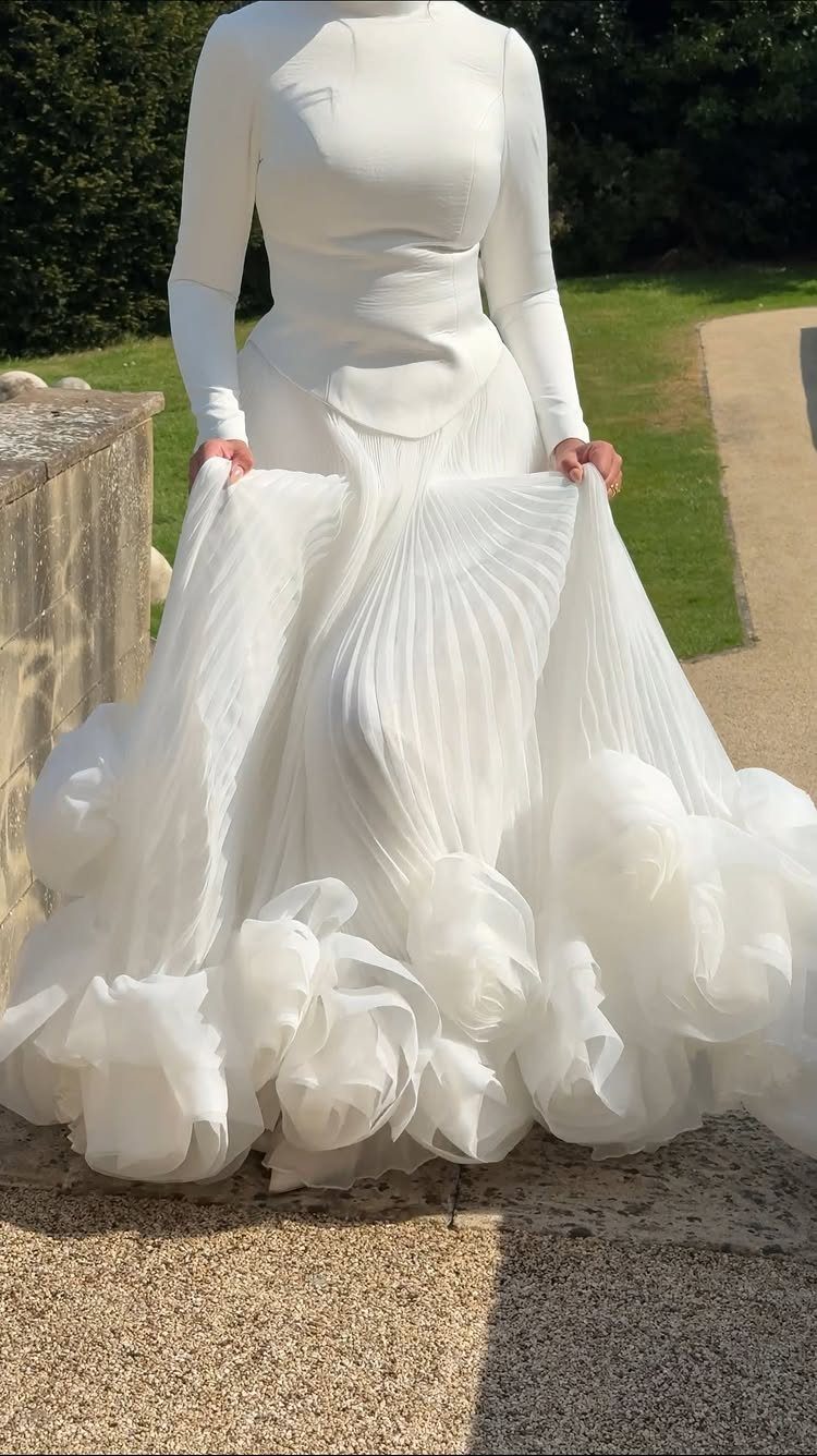 A person in a long-sleeved white wedding dress with a voluminous pleated and ruffled skirt.