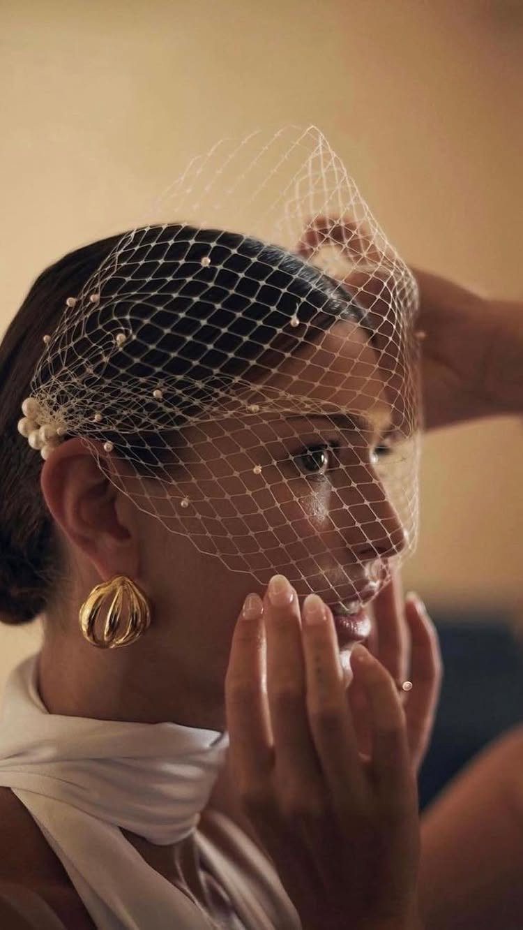 A woman wearing a white pearl-embellished birdcage veil and gold earrings.
