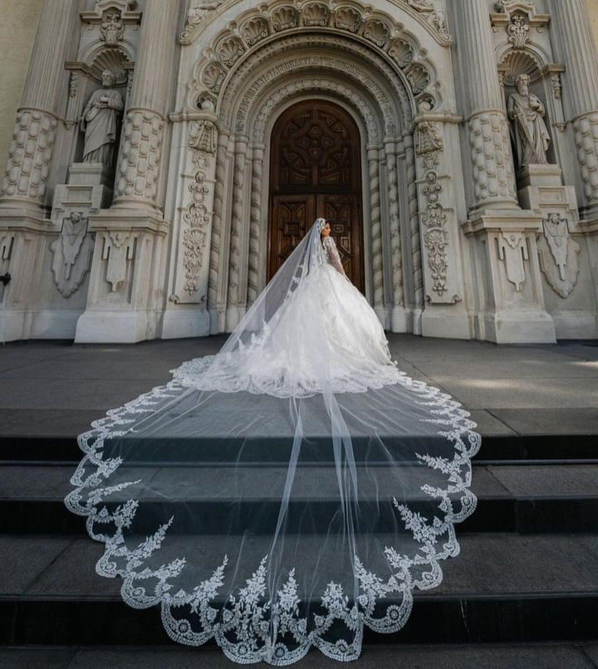 Bride in a white wedding dress wearing a long, lace-edged cathedral veil on cathedral steps.