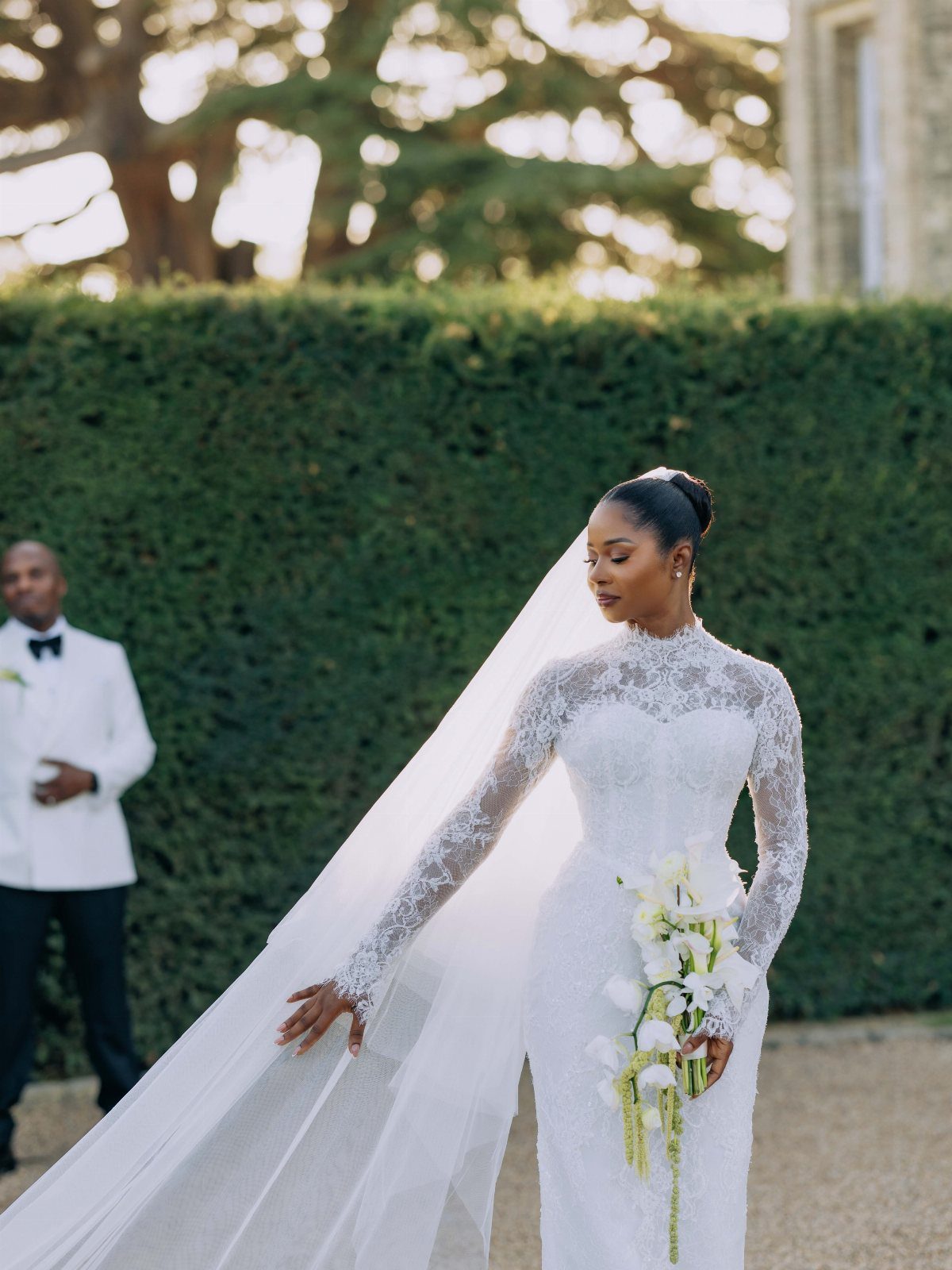 A bride in a lace wedding dress and long veil holding white flowers outdoors.