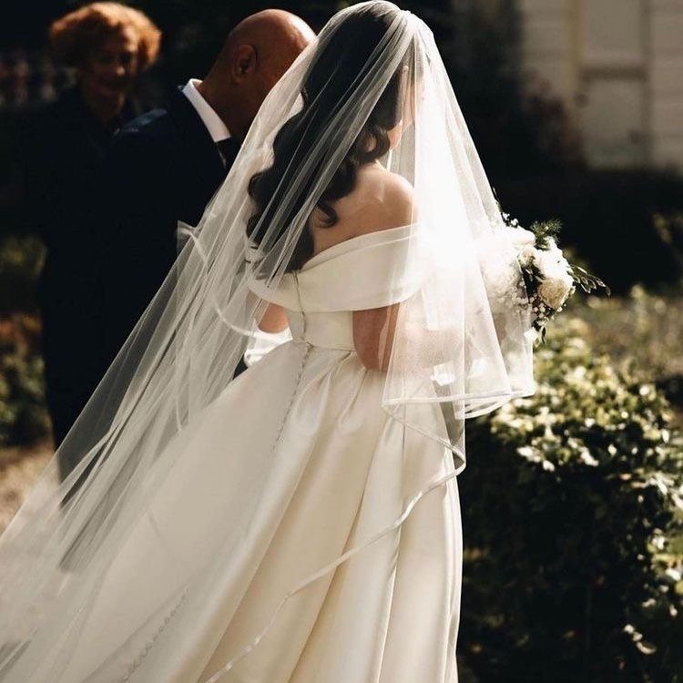 A bride wearing an off-the-shoulder white wedding dress and a long sheer veil holding flowers.