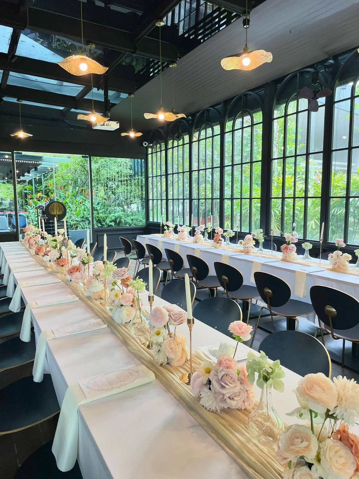 Long rectangular banquet table decorated with pastel flowers and candles in a glass-walled room.