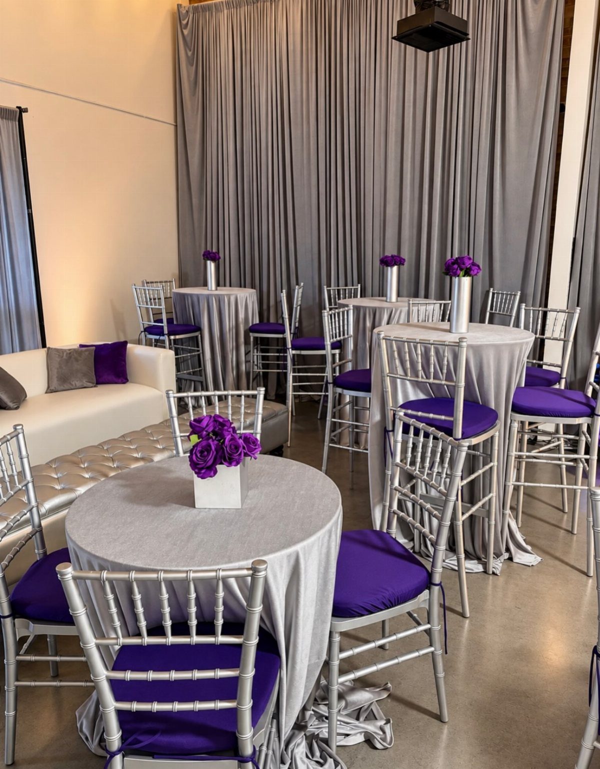Silver cocktail tables with purple cushions and a white sofa in a room with grey curtains.