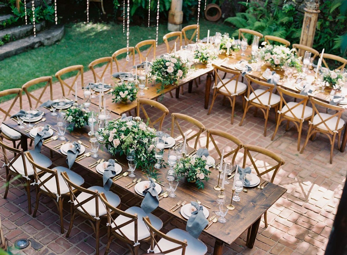 A U-shaped wooden wedding table decorated with white flowers, greenery, and blue napkins on a brick patio.