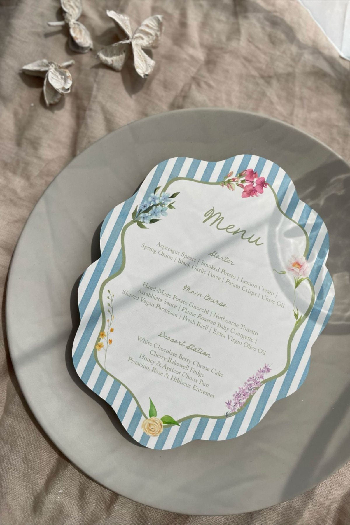 A scalloped menu card with blue stripes and floral illustrations resting on a grey plate.
