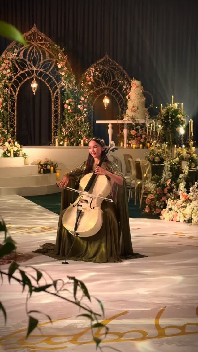 A woman playing a cello in a grand, floral-decorated wedding venue with ornate gold arches.