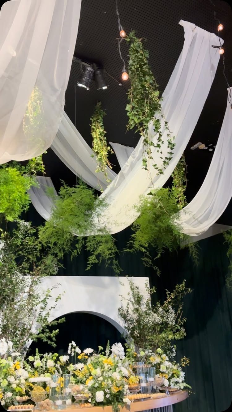 Greenery garlands and white fabric drapes hanging from a dark ceiling above floral arrangements.