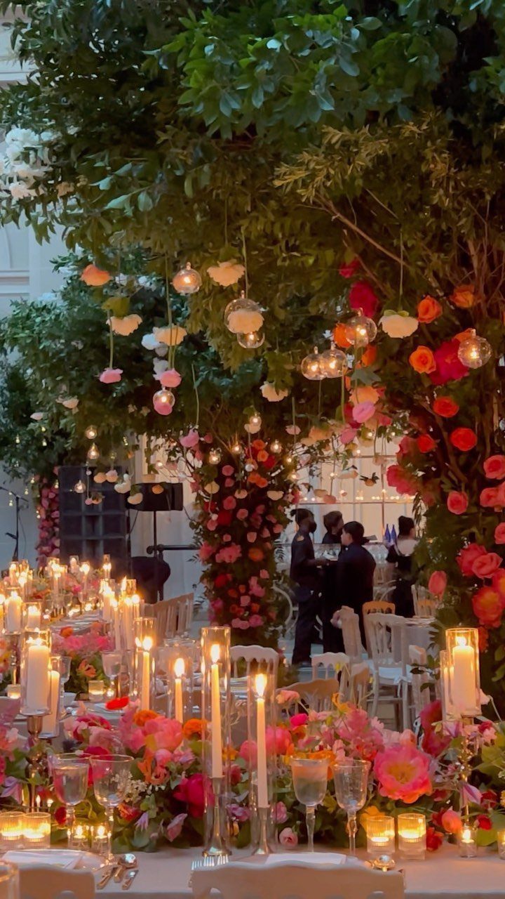 Floral wedding table setting with many lit candles and bright pink flowers under trees.