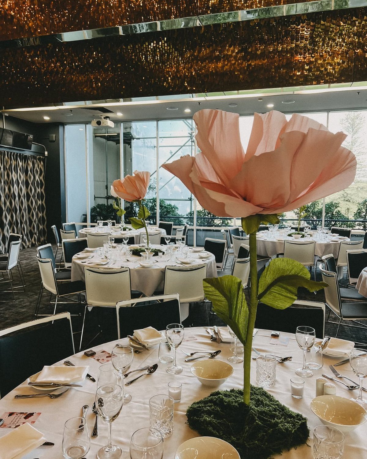 Oversized pink paper flowers acting as whimsical centerpieces on banquet tables in a bright room.