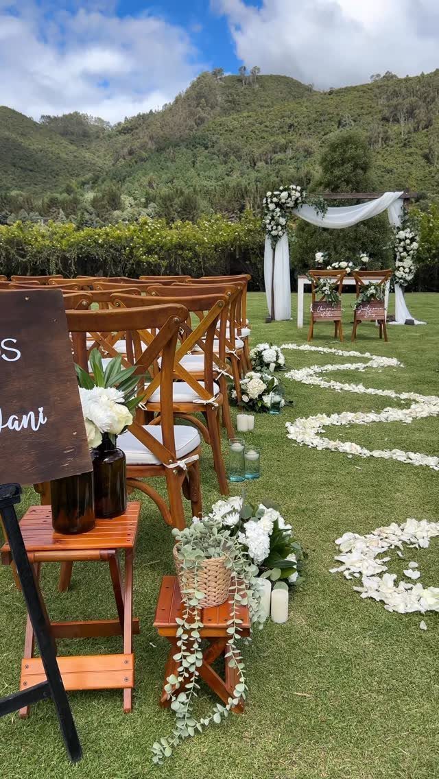 Wooden wedding chairs and white floral arrangements arranged on a grassy lawn near mountains.