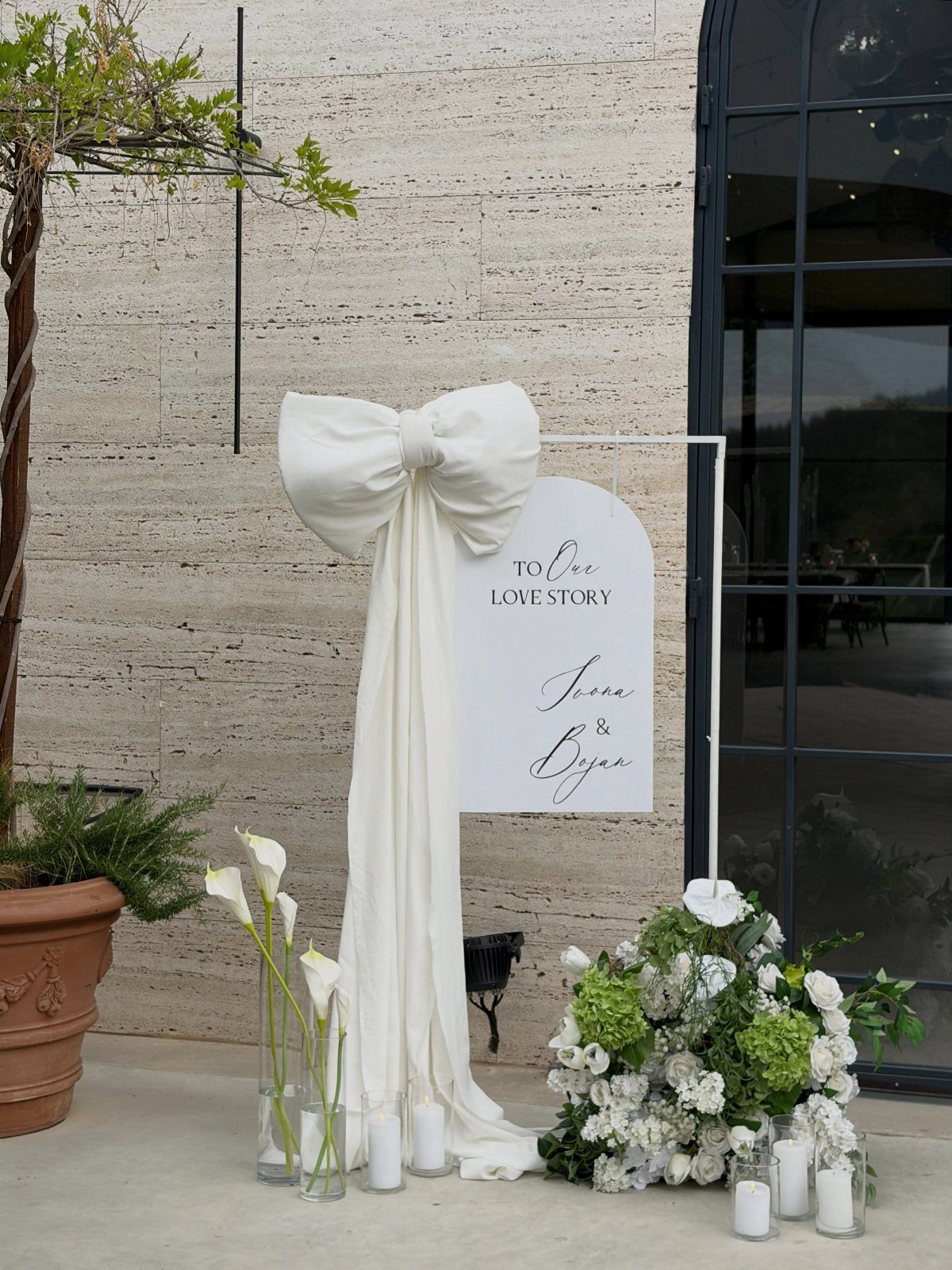 White arched wedding sign with a large fabric bow and white floral arrangements on a patio.