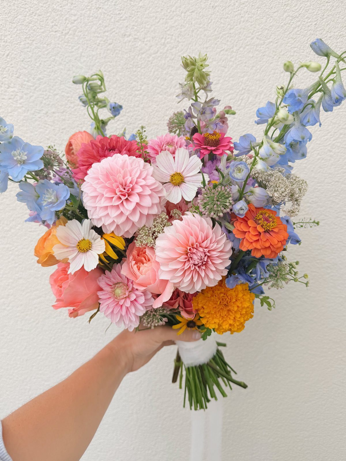 Yellow and Pink Wedding Colors: 6 Ideas for a Sunny Celebration 1 A hand holding a colorful wildflower bouquet with pink dahlias, orange zinnias, and blue flowers.